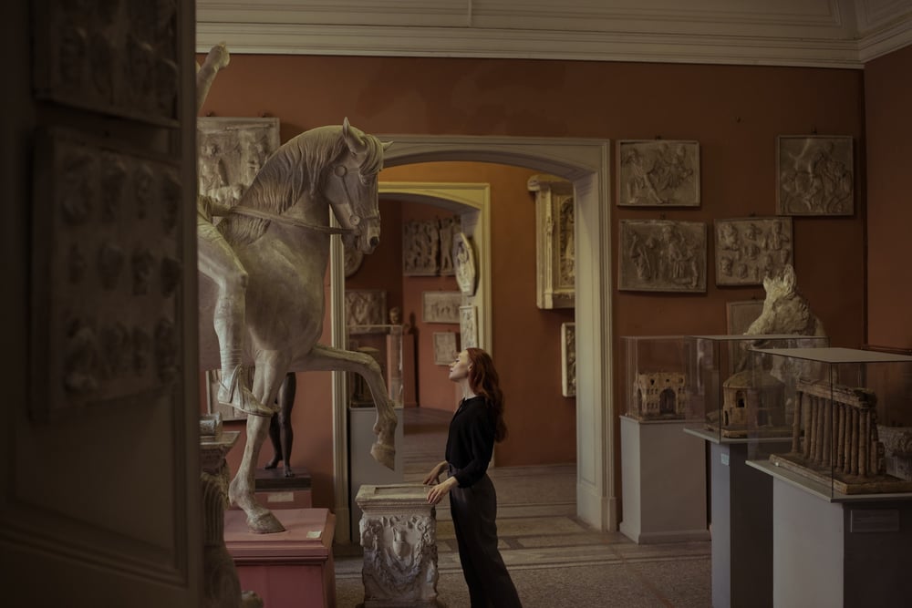 A person with long, red hair examines a horse sculpture in an art museum.