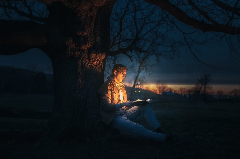 A male-presenting person leans against a tree in the dark, holding a glowing book open.