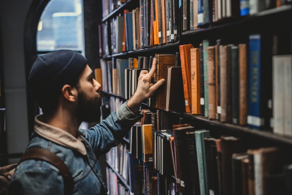 A person with a short beard and a beanie looks at historical books on a library shelf.