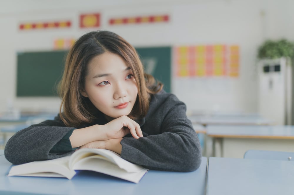 A person sits at a desk with their arms folded over a middle grade book as they star off into the distance.