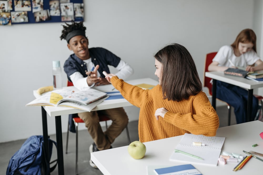 Two middle grade kids in a classroom reaching towards one another with markers.