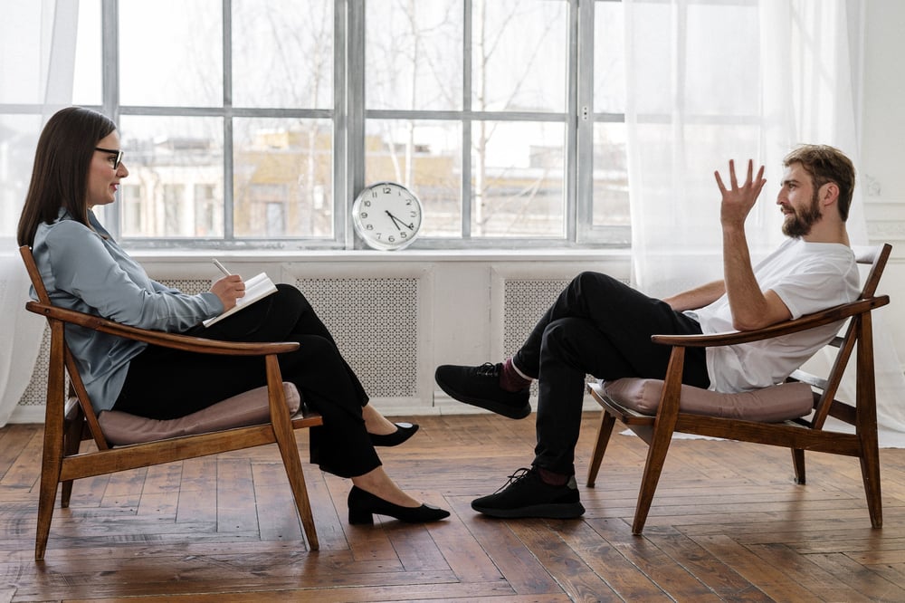 A therapist and a patient sit in chairs opposite one another beside a big window.