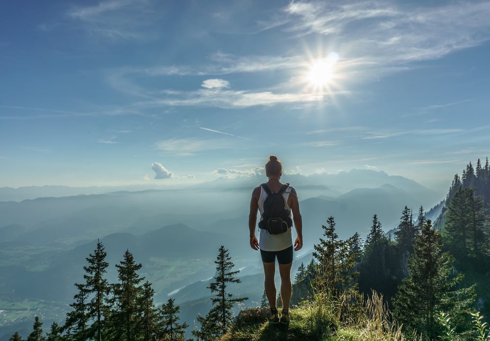 A person wearing a bun and carrying a backpack stands on a mountain overlooking the landscape.