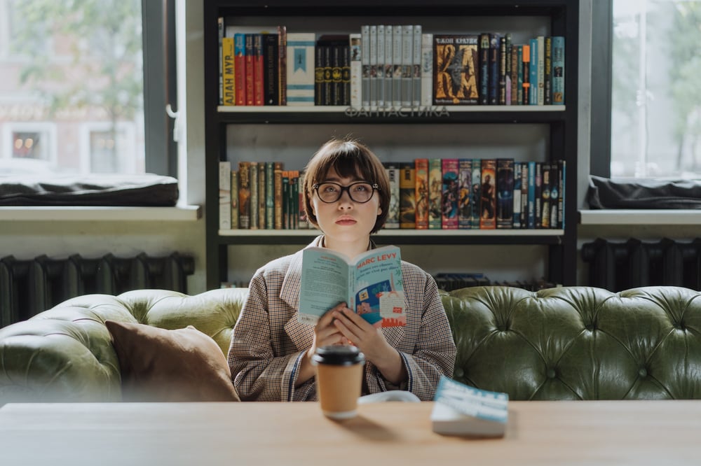 A person with short hair and glasses sits on a green couch holding a book and staring off into the distance.