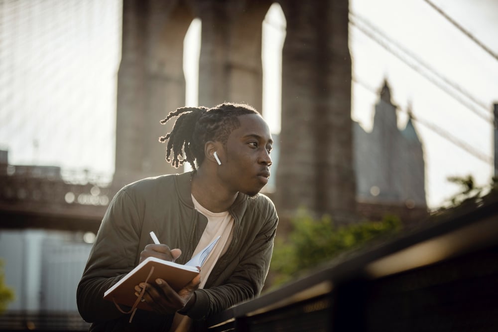 A writer with dreadlocks and air pods leans against a railing outside in front of a bridge while writing in a journal.