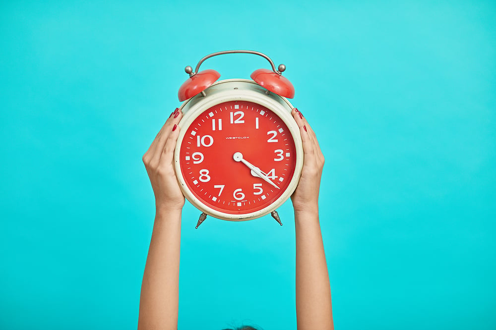 Two hands hold up a bright red analog clock against a blue background.