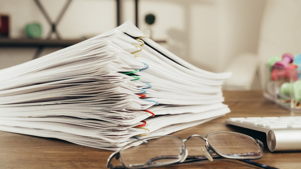 A stack of papers separated with paperclips on a desk beside a pair of glasses and a computer keyboard.