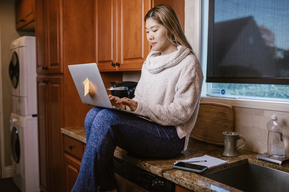 A person with long hair sits on a kitchen counter typing on a laptop.
