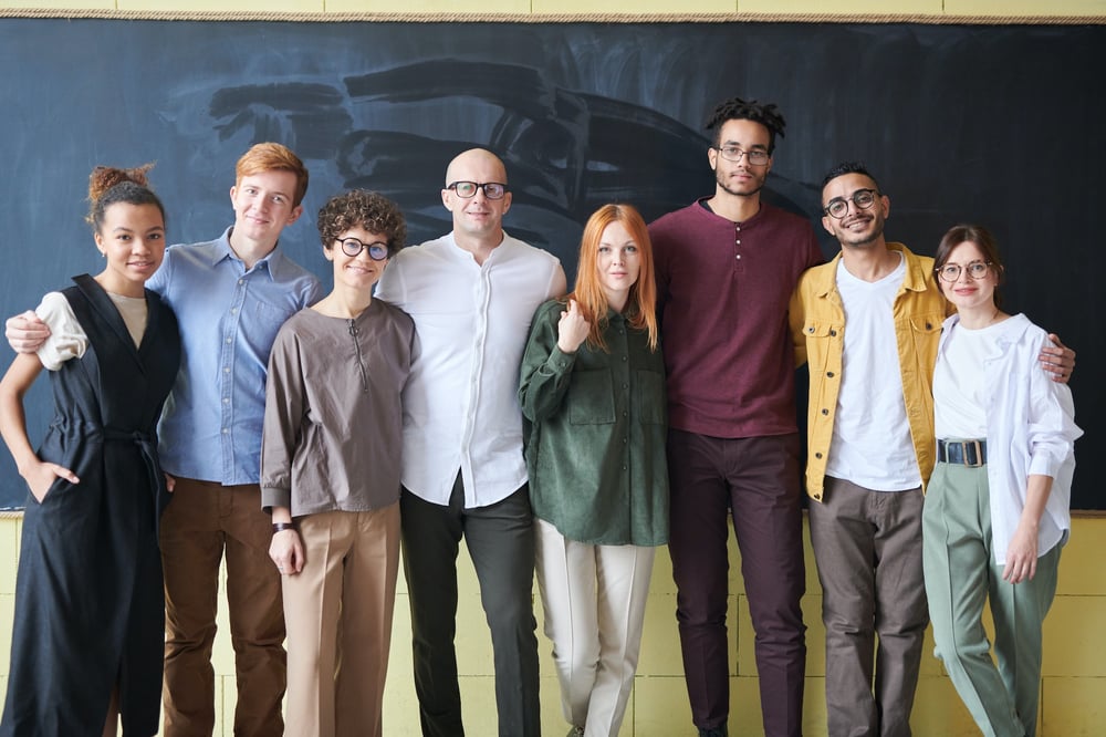 A group of smiling people stand in a line in front of a blackboard.