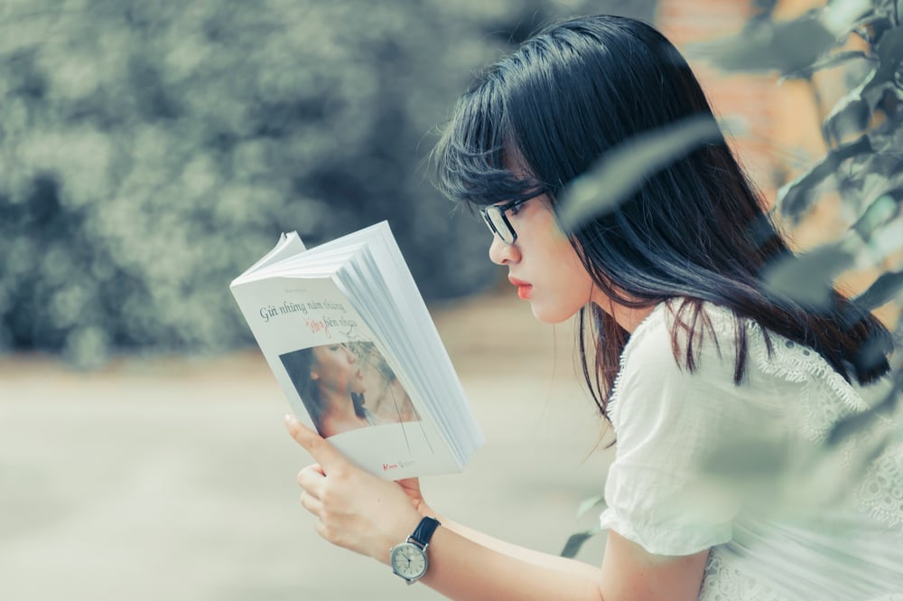 Profile view of a person with long, black hair and glasses engrossed in a great book.