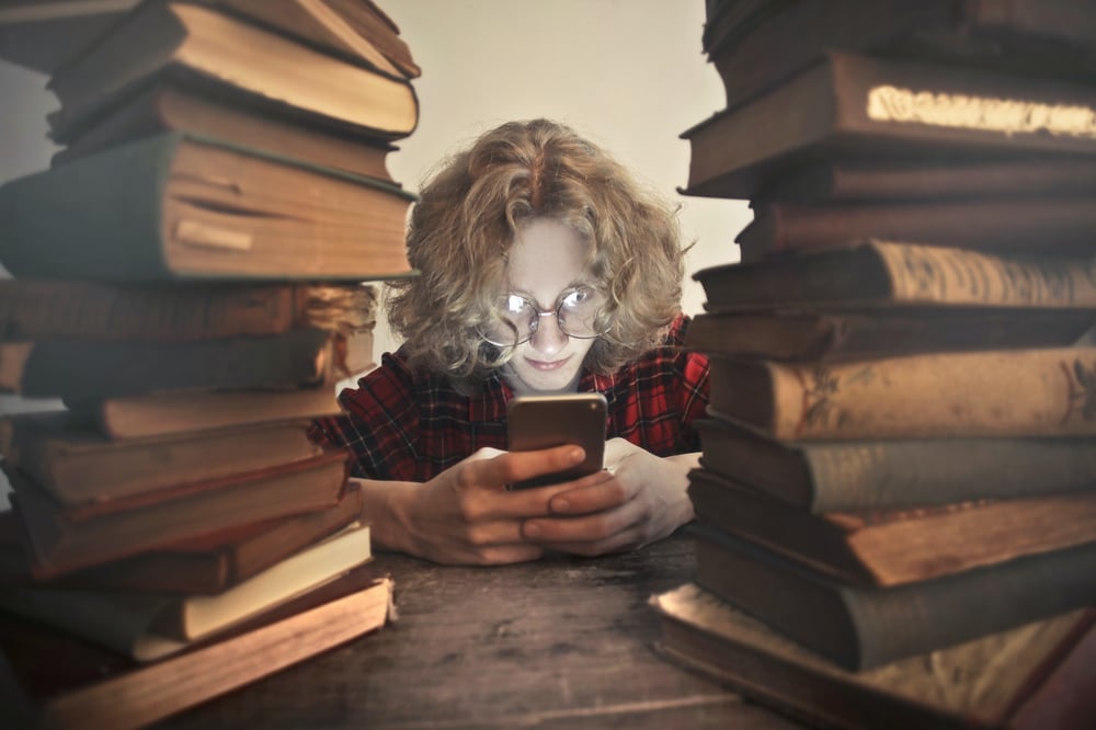 A teenager sits between two stacks of books looking at their phone.