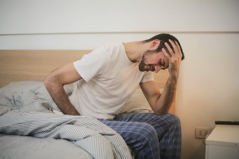 A person sits on the edge of a bed, holding their head in their hand.
