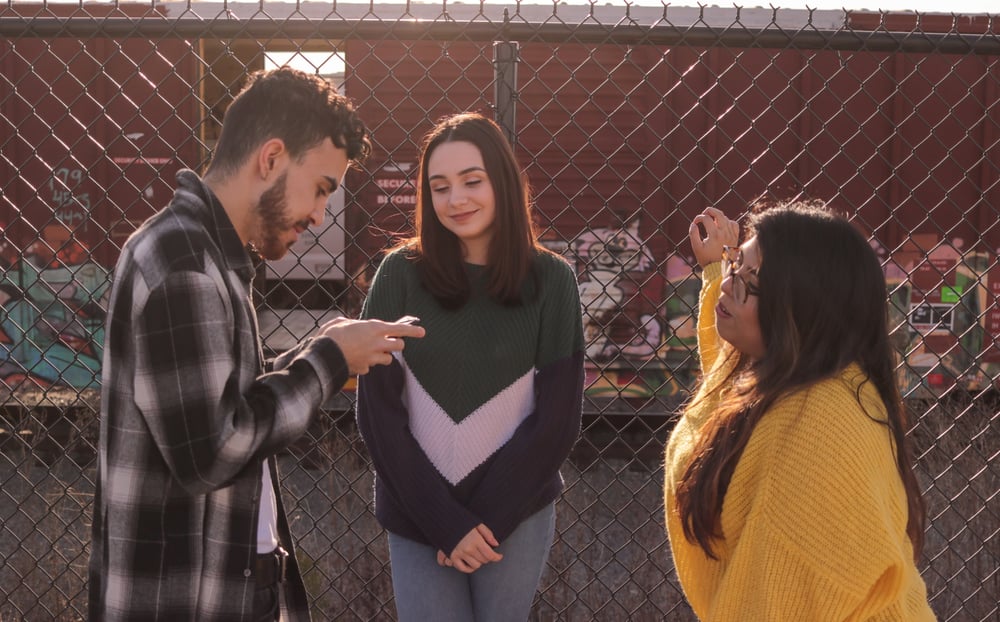 Three teenagers stand by a fence looking at a phone.