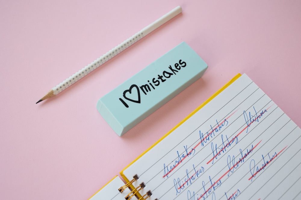 A pencil, a stick eraser that says "I love mistakes," and a notebook containing crossed-out text all against a pink background.