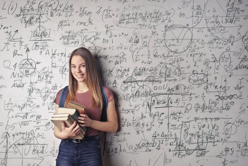 A person with long hair holding books and standing in front of a dry erase board full of math equations.
