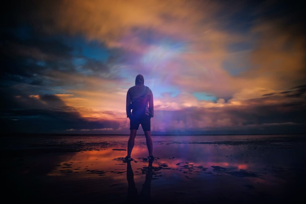 A person stands on a moody beach, watching the sunset.