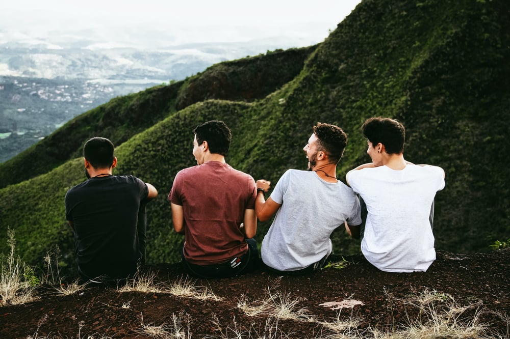 Four friends sit laughing together in a row overlooking green mountains.