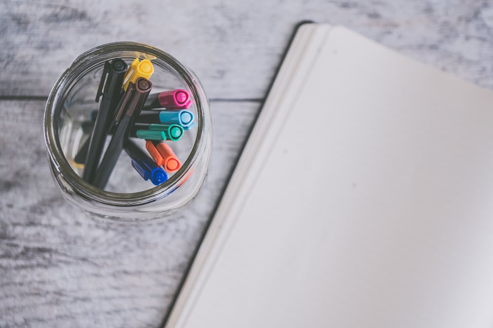 A blank open notebook beside a mason jar containing multi-colored marker pens.