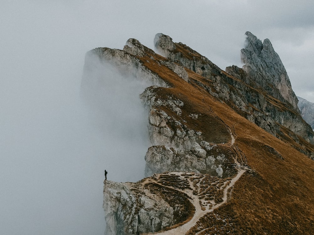 A person stands on the edge of a jagged mountain peak, looking out into mist.
