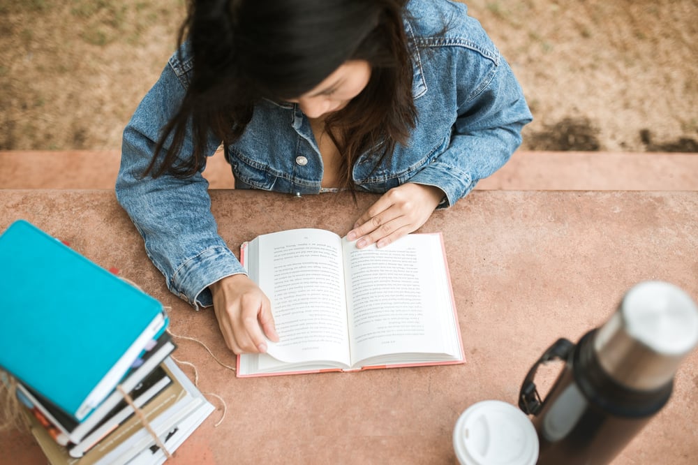 A person in a jean jacket reads a book at an outdoor table next to a stack of books.