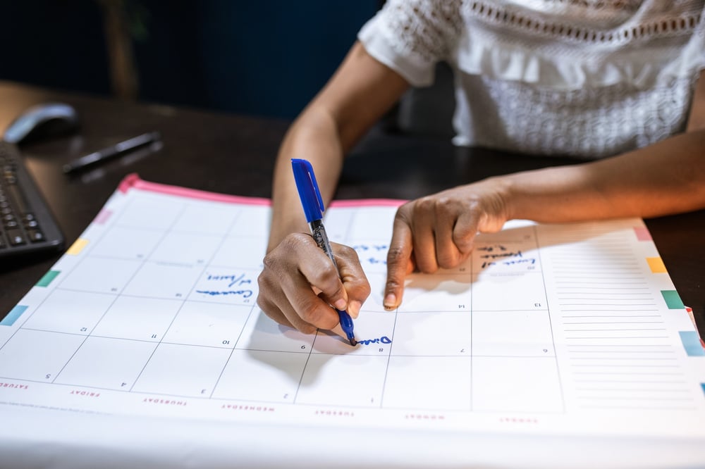 A person in a white blouse writes on a desk-sized calendar.