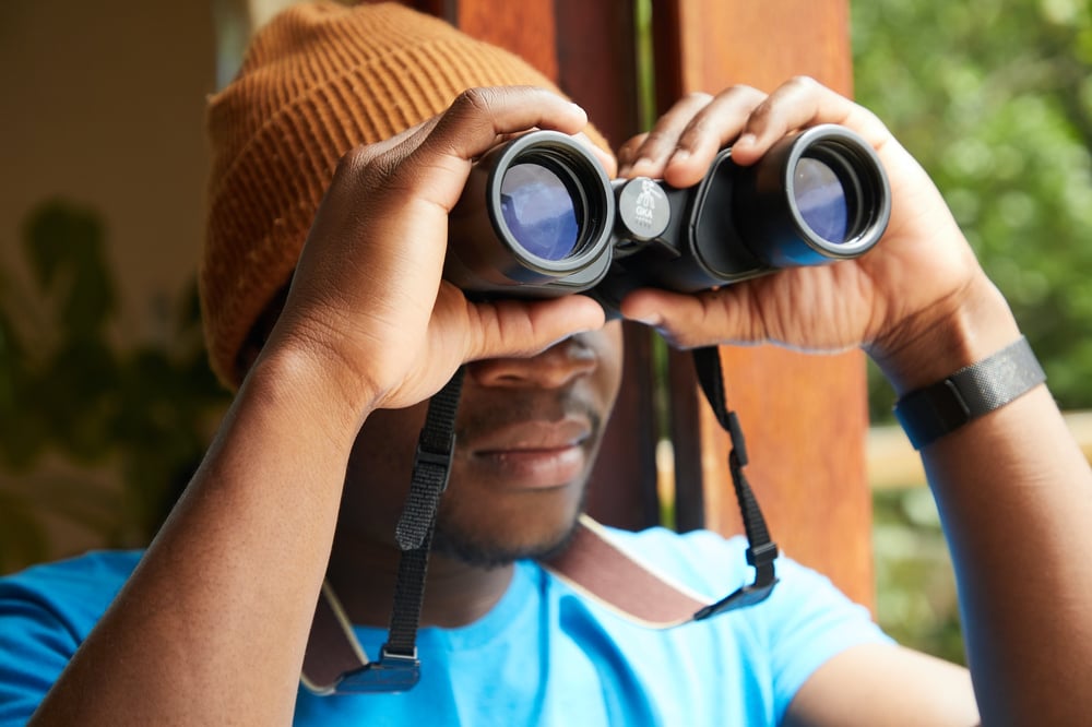 A person wearing an orange beanie stands in doorway and gazes out through binoculars.