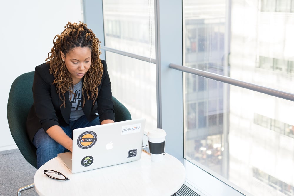 A writer typing on a sticker-covered laptop at a small table beside a high-rise window.