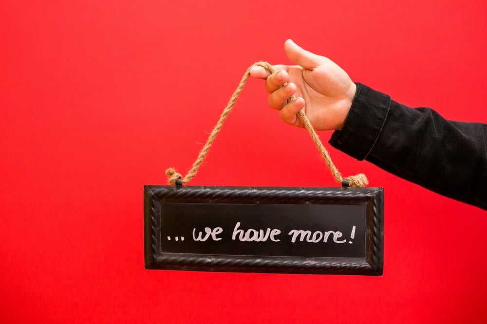 Against a red background, a and holds a small chalkboard sign reading "...we have more!"