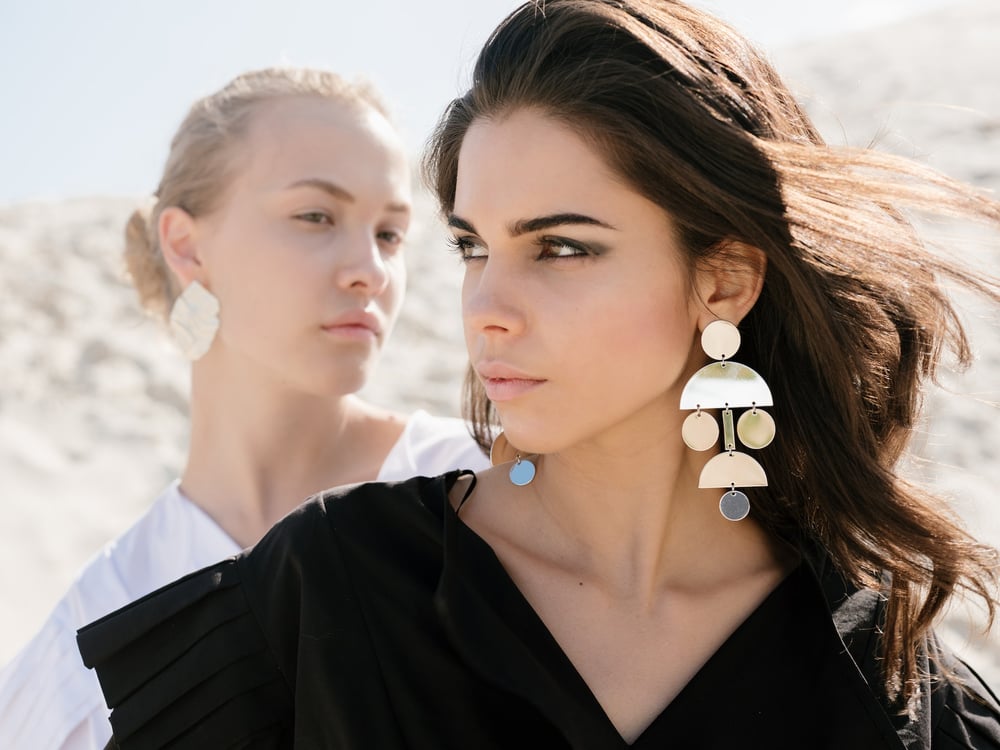 Two people with big, stylish earrings and long hair stand one in front of the other, looking opposite directions.