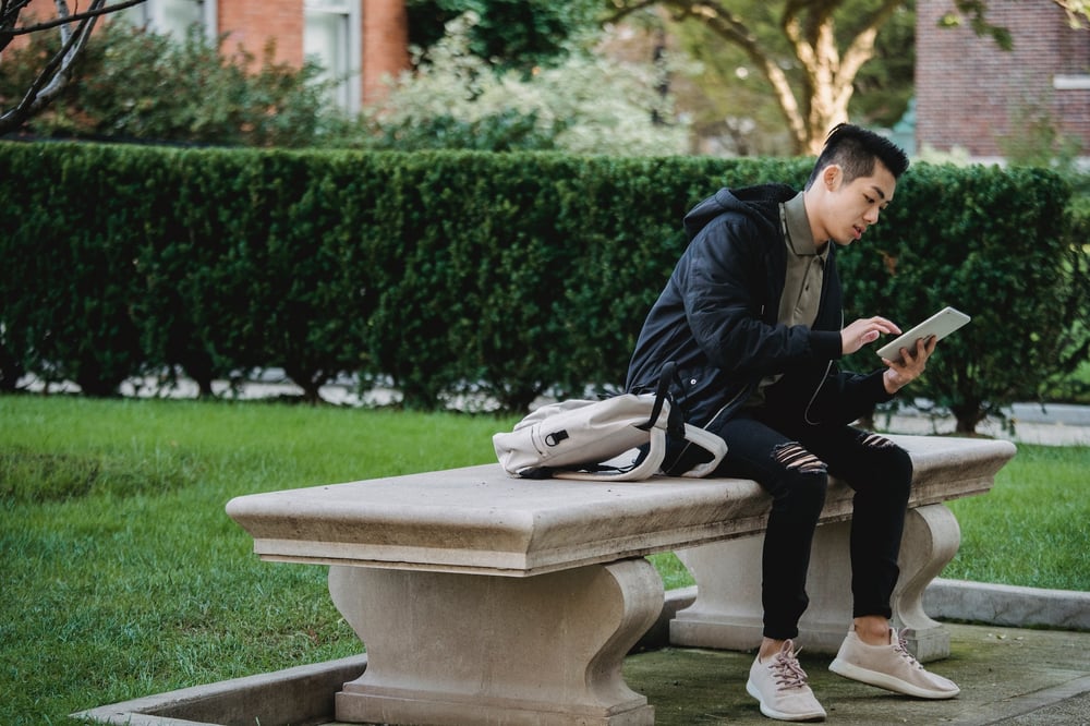 A person sits on a large concrete bench, reading the screen of a tablet.