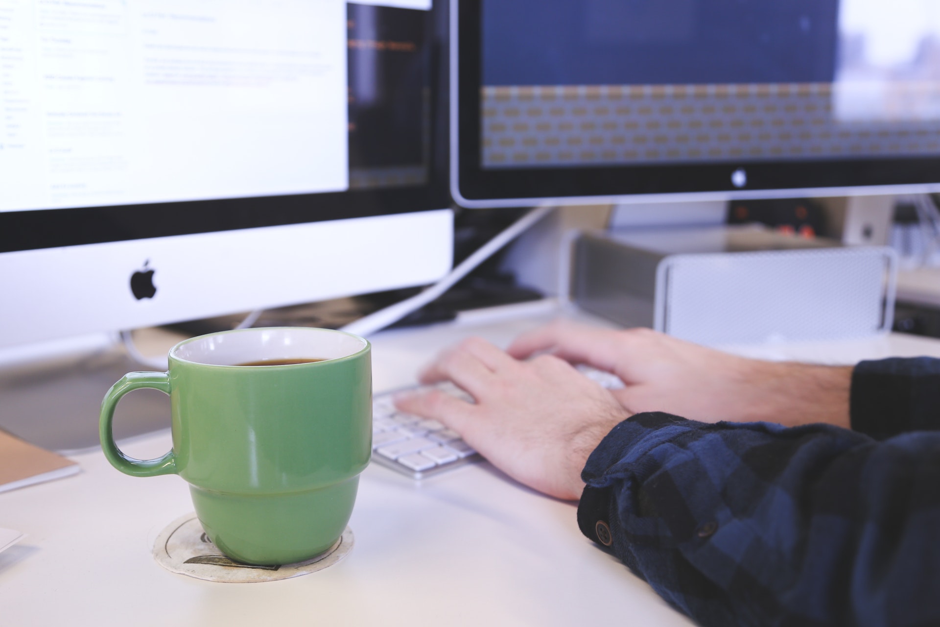 Hands type on a computer keyboard on a white desk. A green coffee cup sits beside the keyboard.