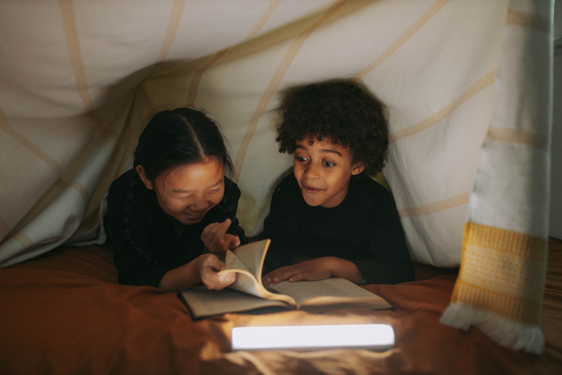 Two children lie on their stomachs under a blanket tent, reading a book together and laughing.