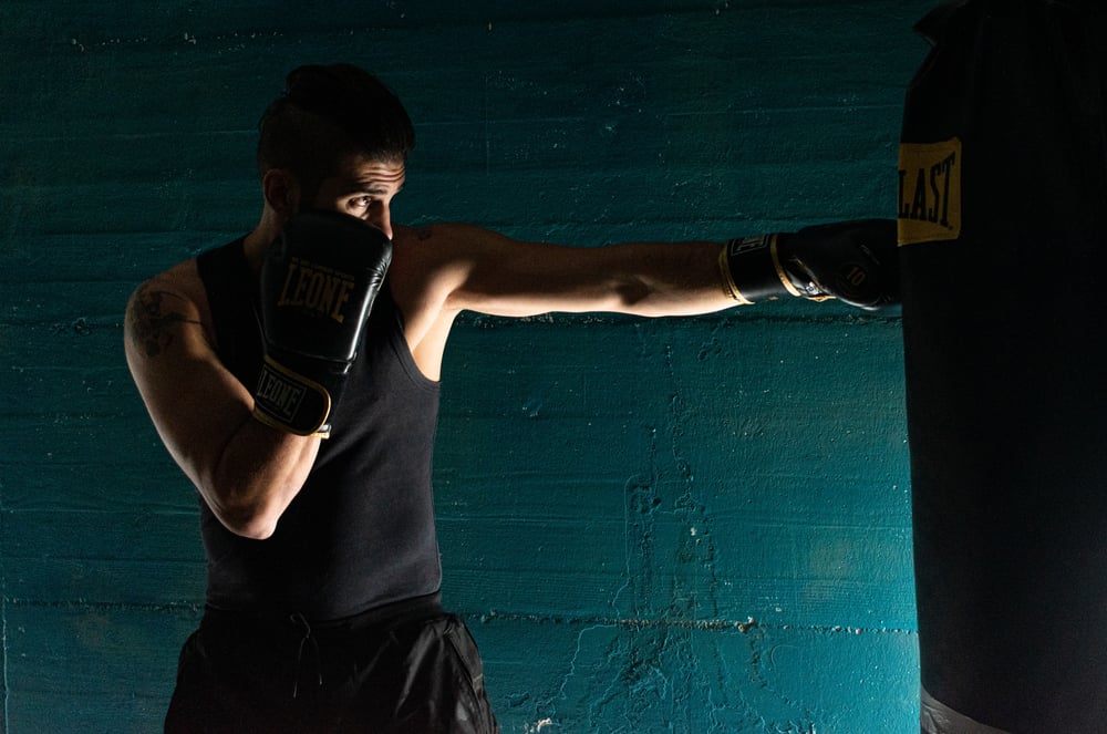 A male-presenting boxer cast in shadow hits a punching bag.