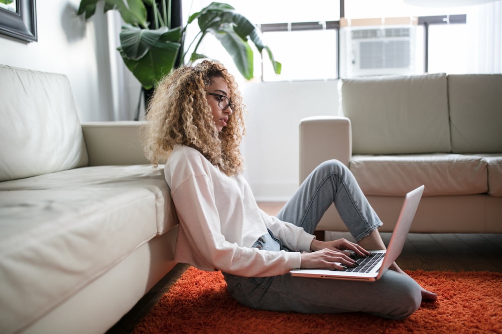 A person with long, curly hair sits on an orange rug beside a white sofa, typing on a laptop computer.