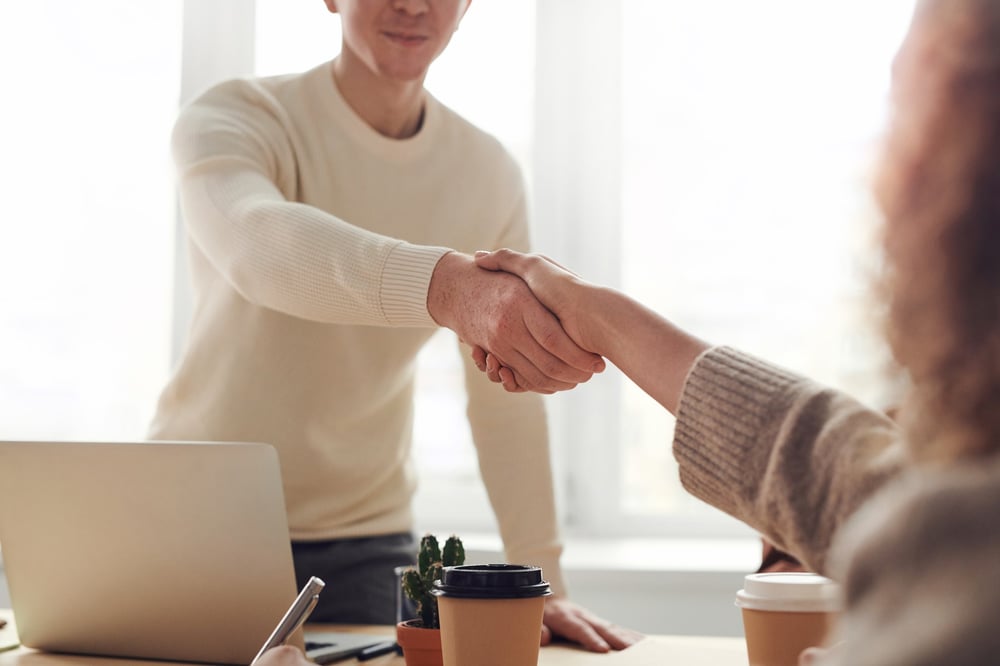 Two people whose faces are not showing shake hands over a desk.