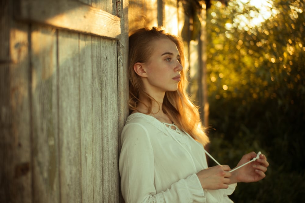 A female presenting person in a white blouse leans against a fence at stares off into the distance.