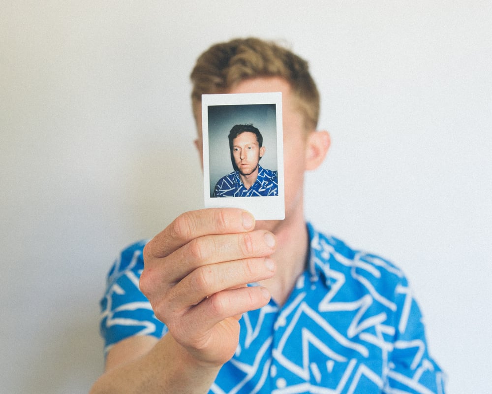 A male-presenting person in a blue shirt holds out a polaroid photo of themselves, covering up their real face.