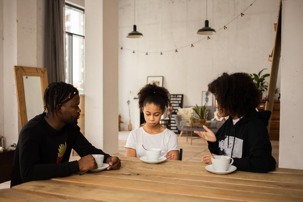 Two adults sit talk to a child at a kitchen table. The child stares down at a large teacup.