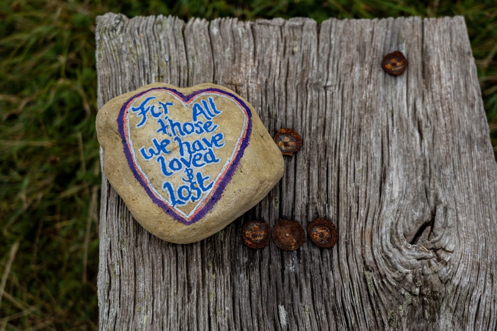 A painted stone reading "For all those we have loved & lost" sits on top of a wood bench.
