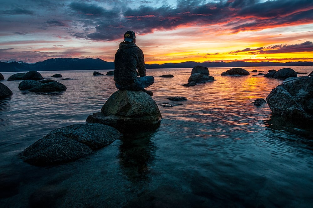 A male-presenting person in a hoodie and baseball cap sits on a rock in the ocean watching the sun set.