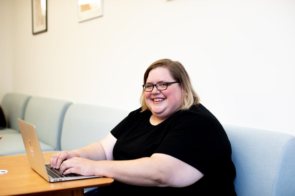 A female-presenting person with glasses sits in a booth and smiles while typing on a laptop.