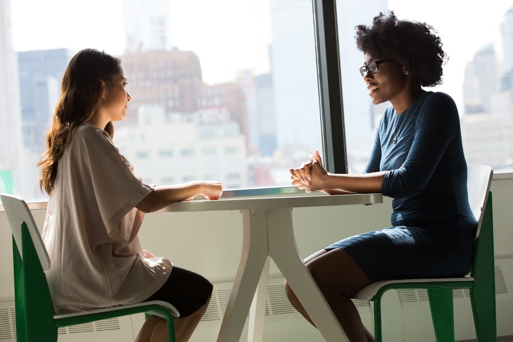 Two female-presenting people have a conversation over a table in front of a window overlooking a city.
