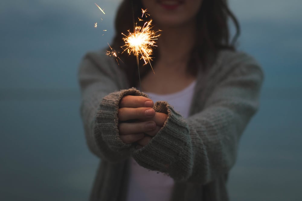 A person in a gray sweater holds out a lit sparkler.