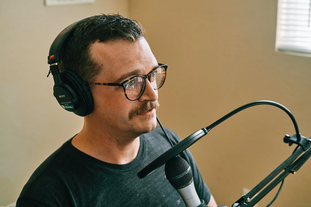 An audiobook narrator wearing headphones stands in front of a microphone.