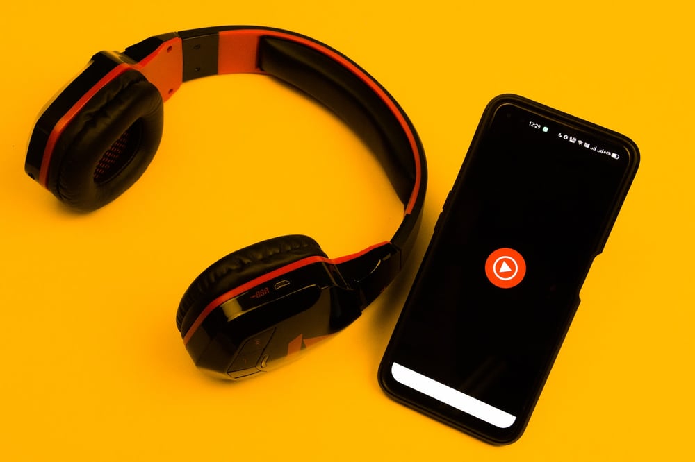 Red and black headphones on a yellow background beside a black smartphone with a play button in the center of the screen.o