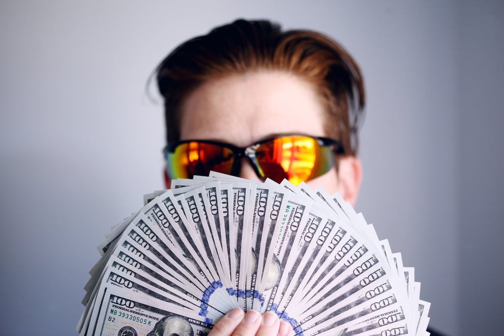 A person with short hair and reflective sunglasses fans a bunch of $100 bills in front of their face.
