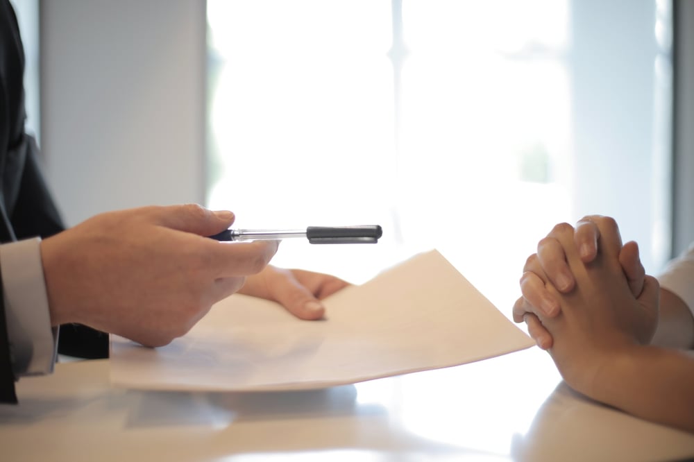 A pair of hands pass a contract and a pen to a folded pair of hands.