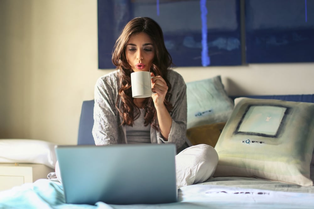 A writer sits on a bed with a laptop, blowing on a cup of coffee.