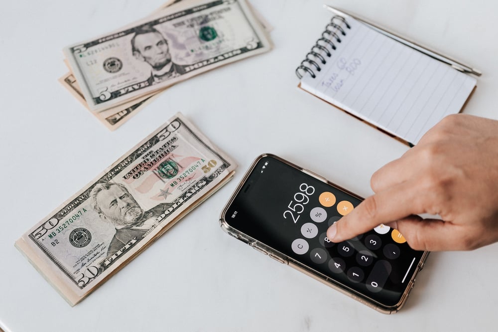 A hand pushes numbers on a smartphone's calculator app. Stacks of money and a notebook sit nearby.