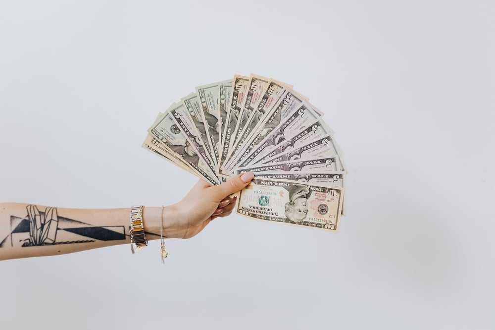 An arm with tattoos and bracelets holds a bunch of cash fanned out.